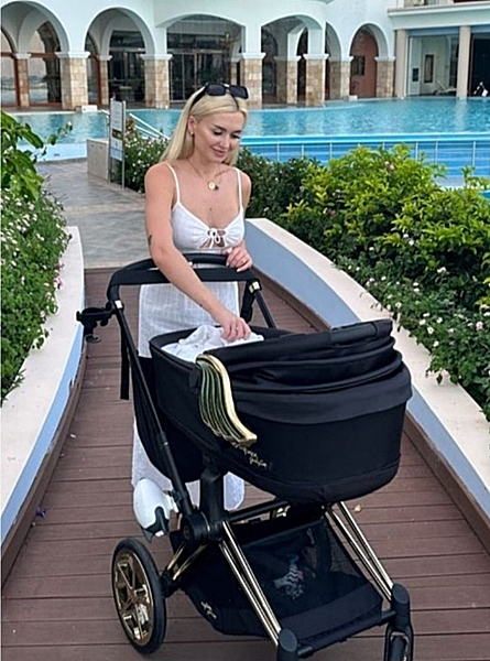 Influencerka wersow spaceruje drewnianą alejką obok basenu, prowadząc elegancki czarny wózek dziecięcy cybex ze złotymi detalami. W tle widoczne krzewy, kwiaty oraz budynek z arkadami i błękitną wodą w basenie. Kobieta ma blond i włosy, na głowie okulary przeciwsłoneczne i delikatną biżuterią.